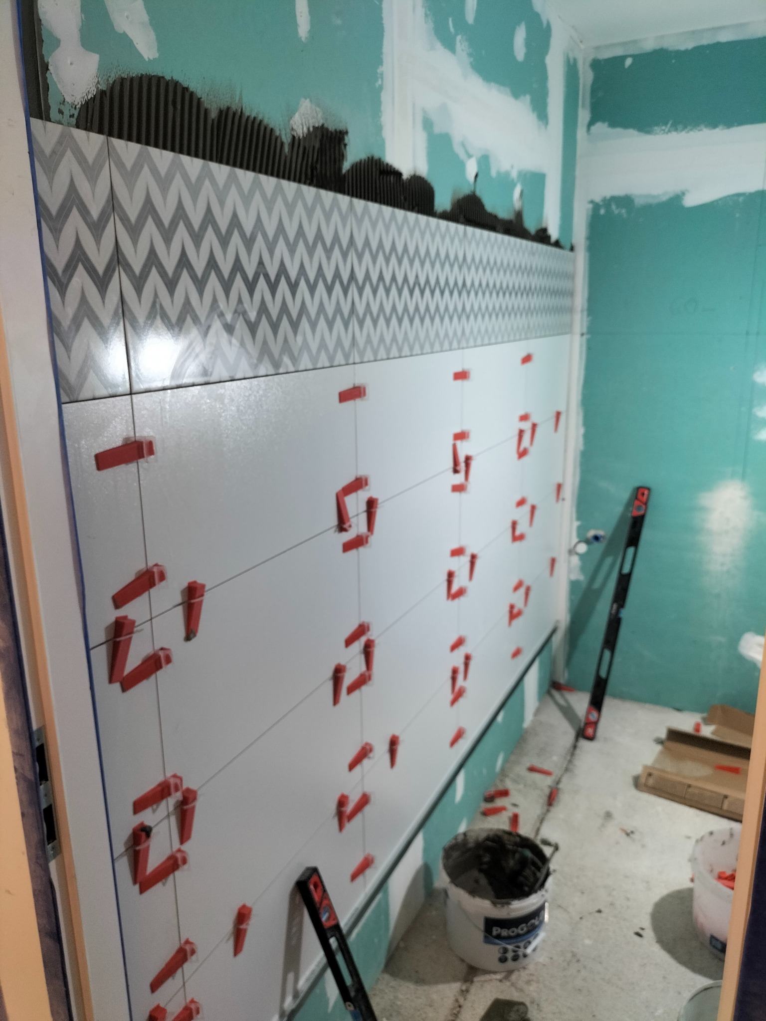 Układanie płytek ściennych w łazience, widoczne poziomice, wiaderko z klejem i elementy dystansowe na świeżo położonych płytkach z wzorem chevron.