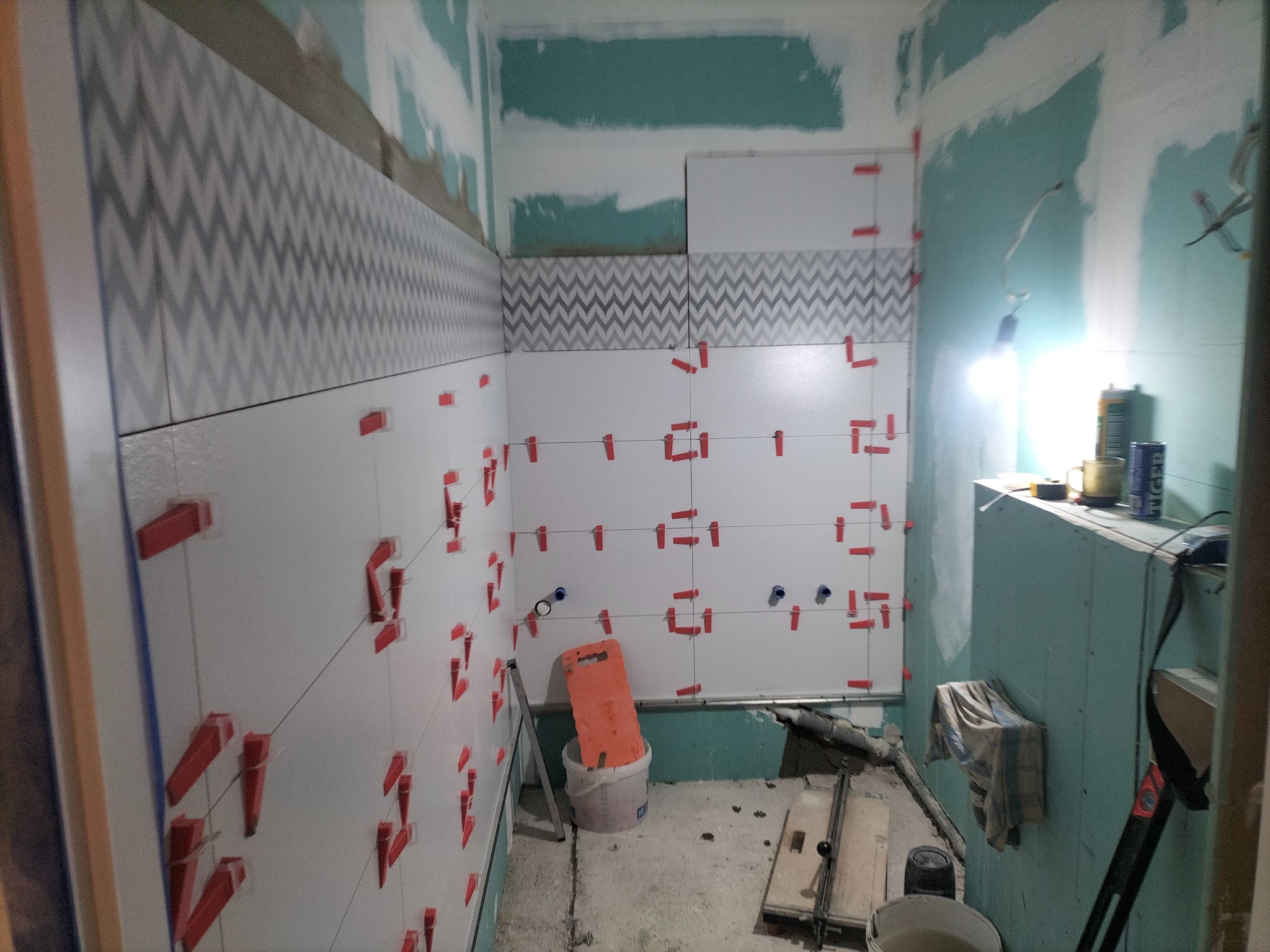 Montaż białych płytek z szarym zygzakowatym wzorem w łazience, widoczne czerwone krzyżyki dystansowe, przygotowane ściany i narzędzia.
