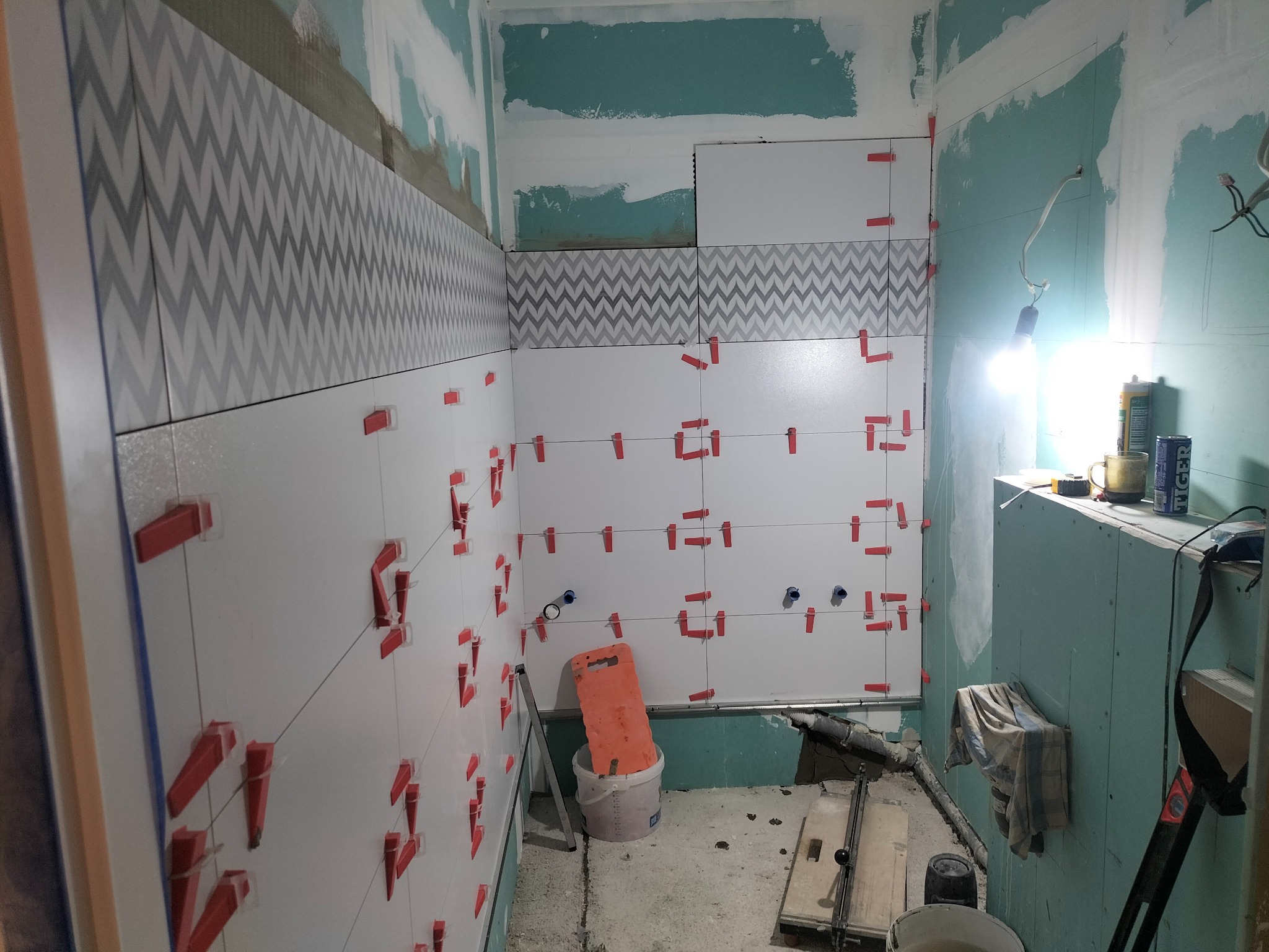 Montaż płytek w łazience, widoczne białe i szare płytki z wzorem chevron, system poziomowania płytek z czerwonymi klipsami, przygotowane otwory na instalacje, narzędzia i materiały budowlane w tle.