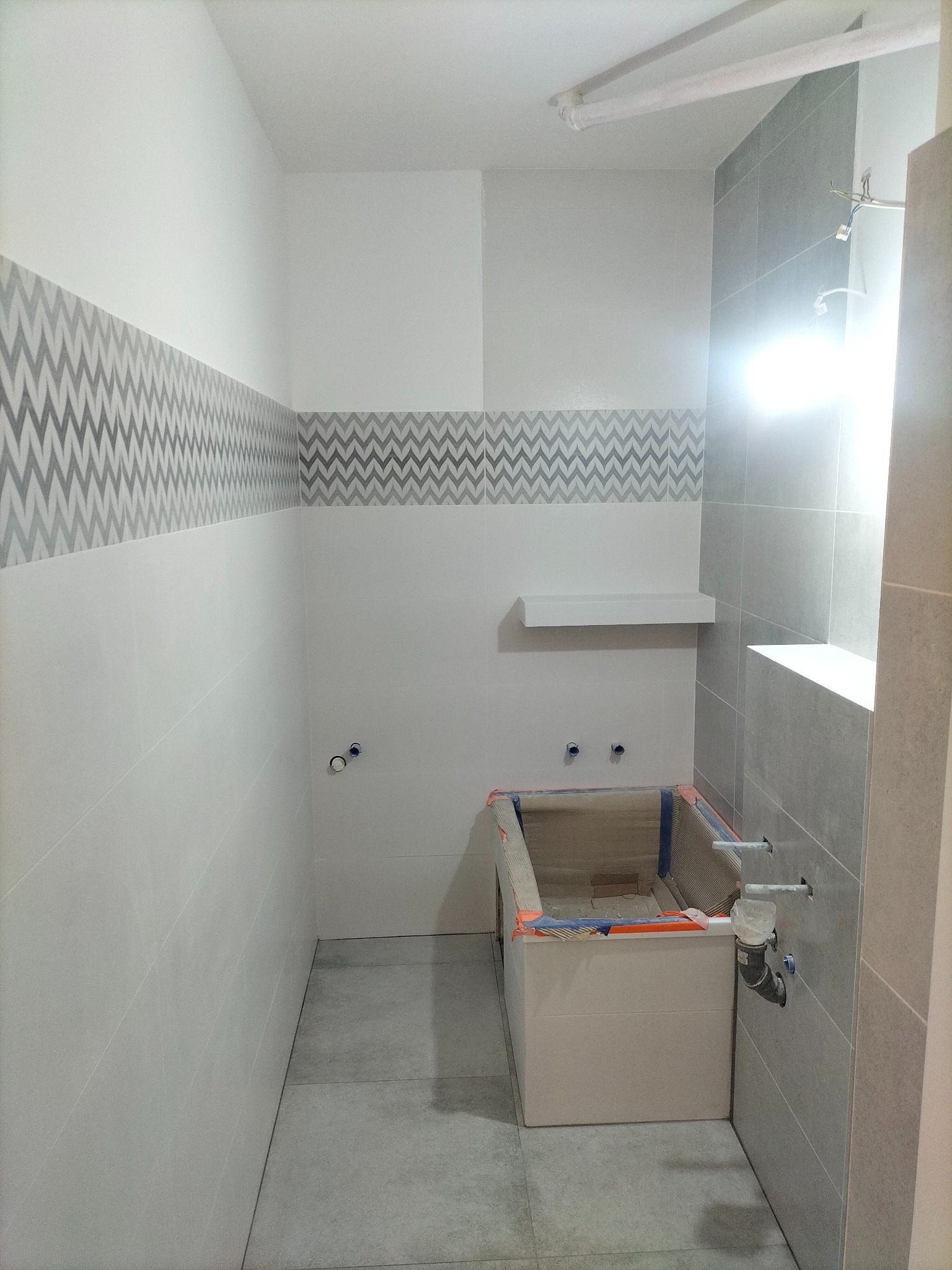 Wnętrze łazienki w trakcie remontu, widoczna obudowana wanna przygotowana do wykończenia, szare płytki na podłodze i ścianach z dekoracyjnym pasem wzorów, instalacja wodna w trakcie podłączania.