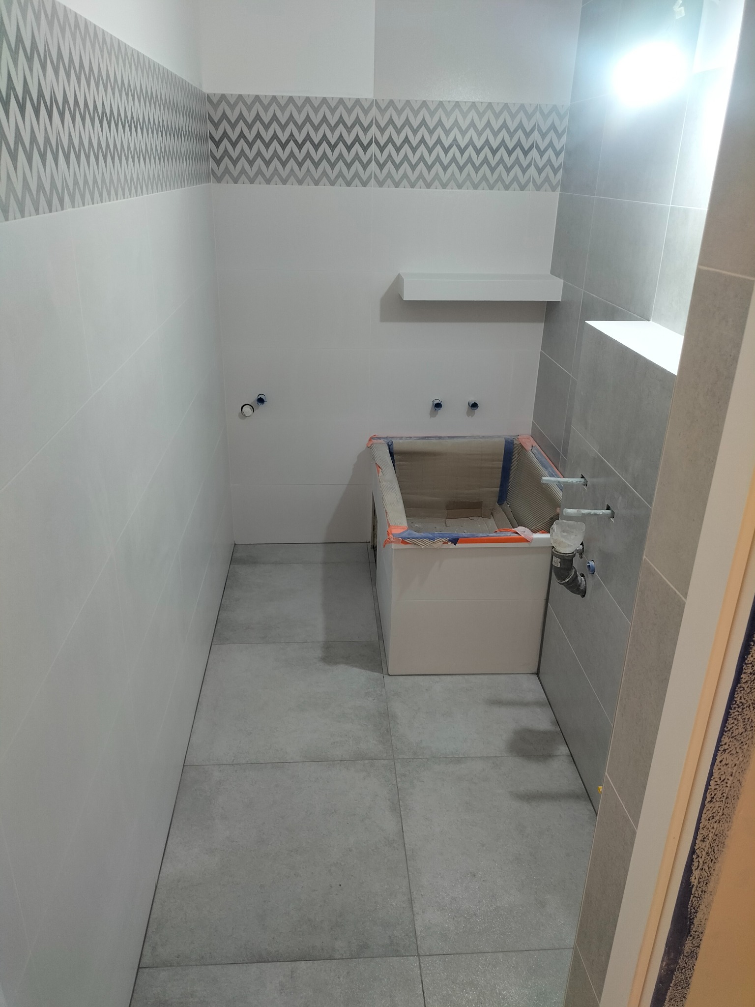 Wnętrze łazienki w trakcie remontu, z widoczną obudową wanny zabezpieczoną folią, szare płytki na podłodze i ścianach, dekoracyjny pas płytek z wzorem zygzaków.