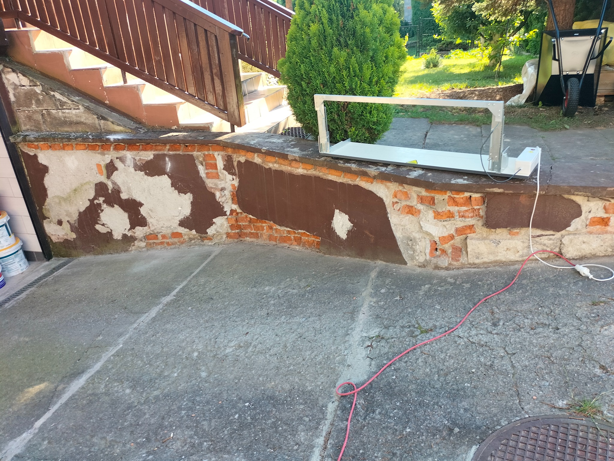 Przygotowanie do prac brukarskich: poziomica laserowa ustawiona na nierównym murku z cegły, widoczne ślady napraw i ubytki, w tle schody i zieleń.