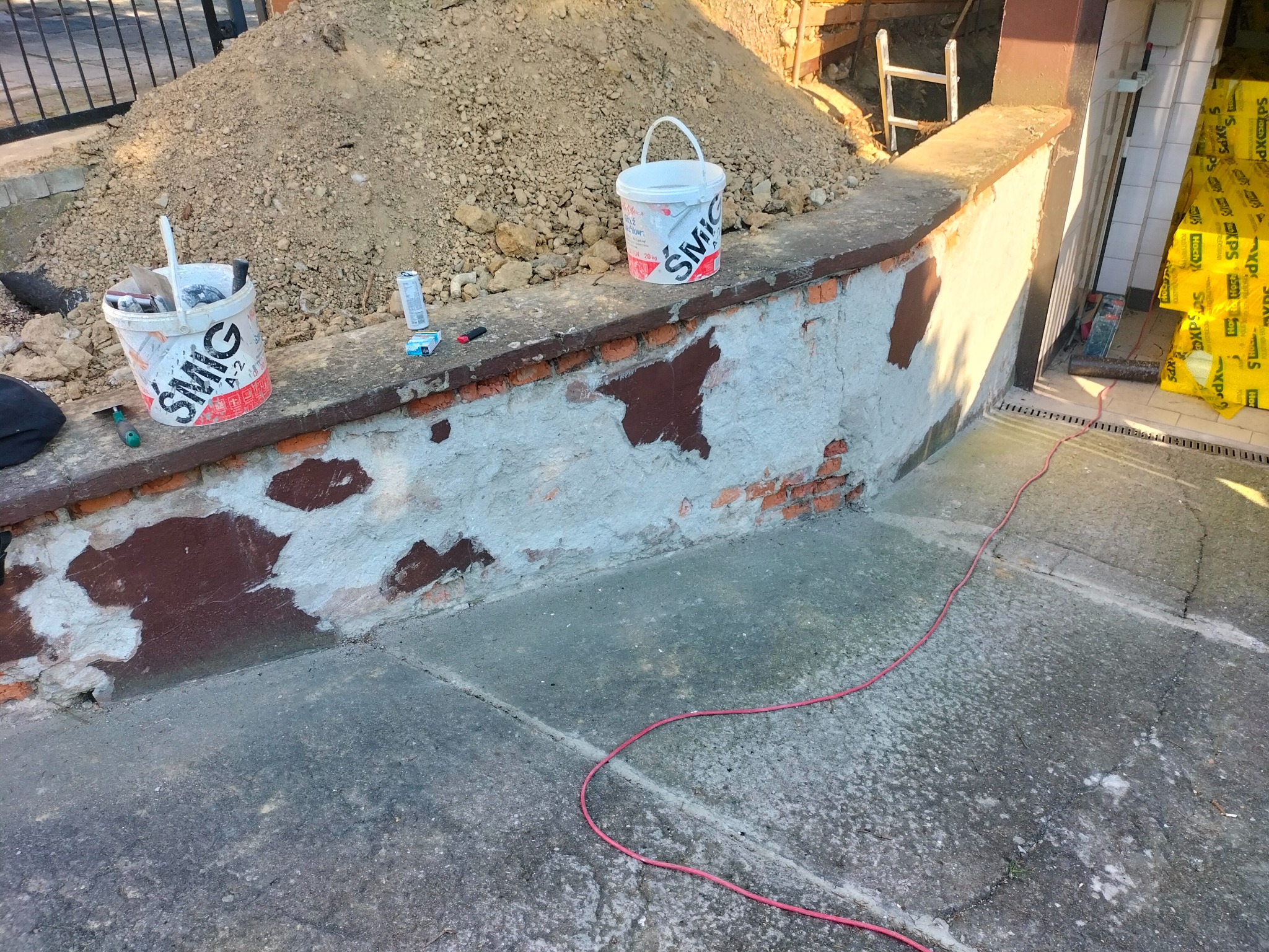 Przygotowania do prac murarskich: dwa wiadra z zaprawą SMIG stoją na murku z częściowo odnowioną elewacją, w tle sterta piasku, narzędzia i materiały izolacyjne.