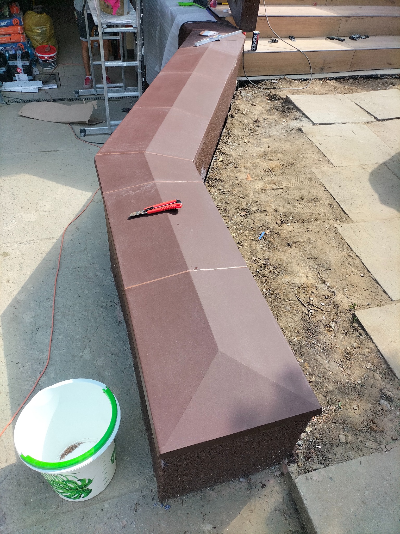 Brązowa, kanciasta obudowa z kostki przy schodach, w trakcie montażu, z nożykiem na wierzchu, obok wiadro i fragment ułożonej kostki brukowej.