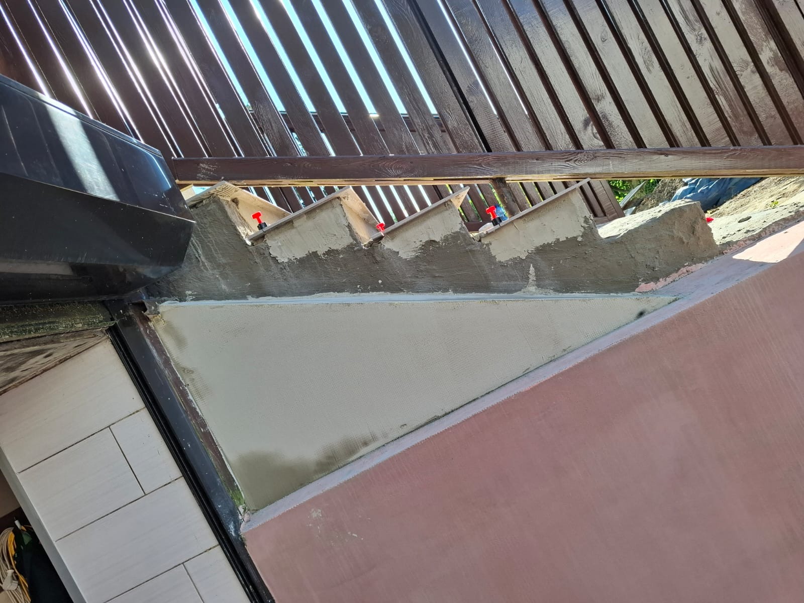 Fragment dachu z widocznymi elementami konstrukcyjnymi podczas prac wykończeniowych, z użyciem poziomujących klipsów dystansowych, na tle drewnianej konstrukcji pergoli.