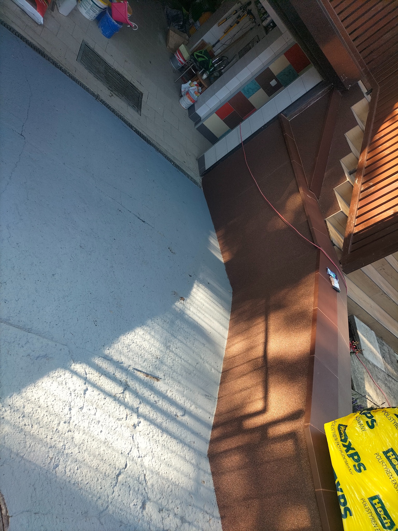 Widok z góry na fragment dachu z brązową posypką bitumiczną, jasnoszarą powierzchnię przypominającą beton, żółtą folię z napisem XPS oraz elementy drewnianej konstrukcji dachu w słoneczny dzień.