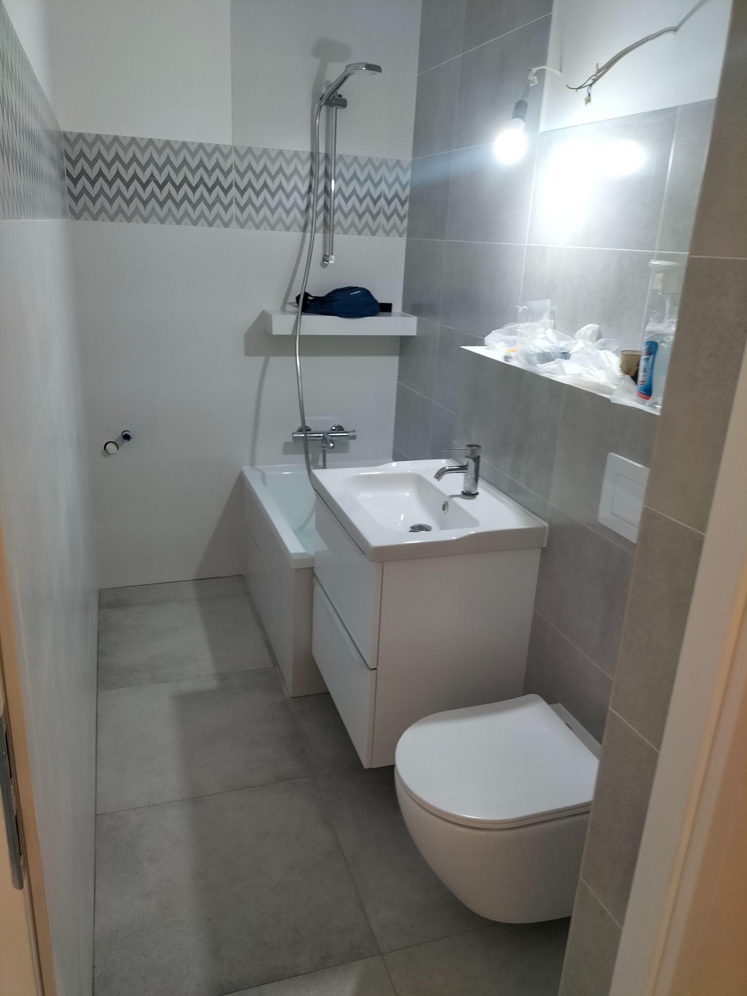 Nowoczesna łazienka z szaro-białą kolorystyką, widoczne: wanna z prysznicem, umywalka z szafką, toaleta podwieszana, szare płytki na ścianach i podłodze, oraz dekoracyjny pas z motywem zygzaków.