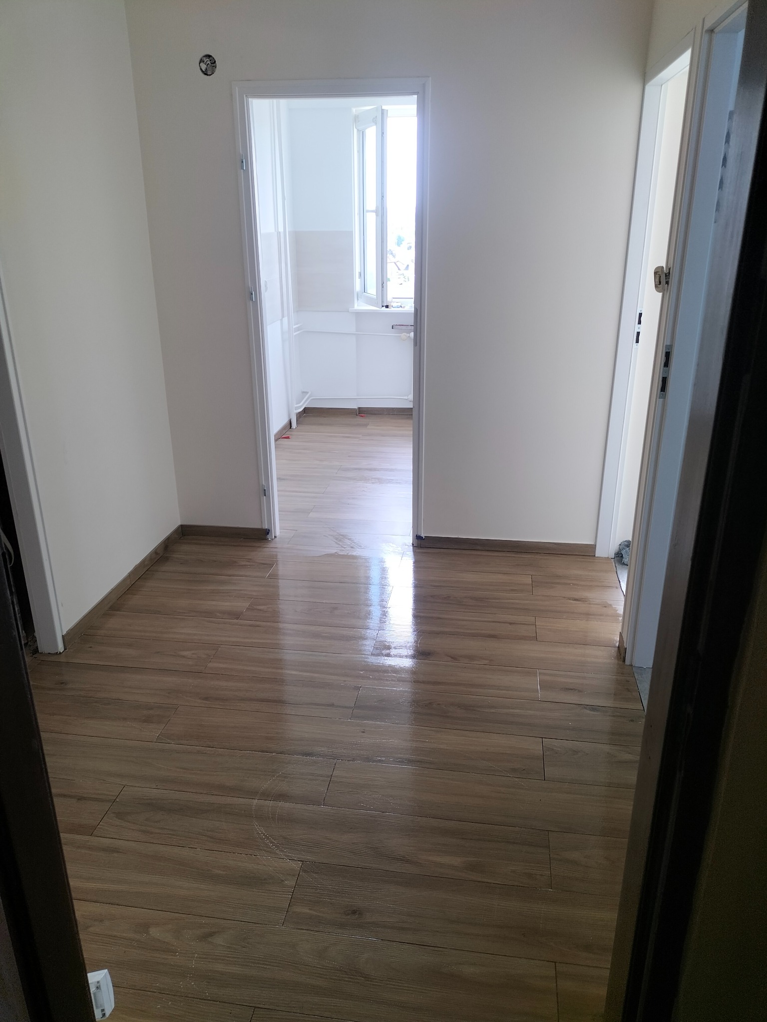 Ułożona podłoga z płytek imitujących drewno w korytarzu, widok na białe ściany i otwarte drzwi do jasnego pomieszczenia