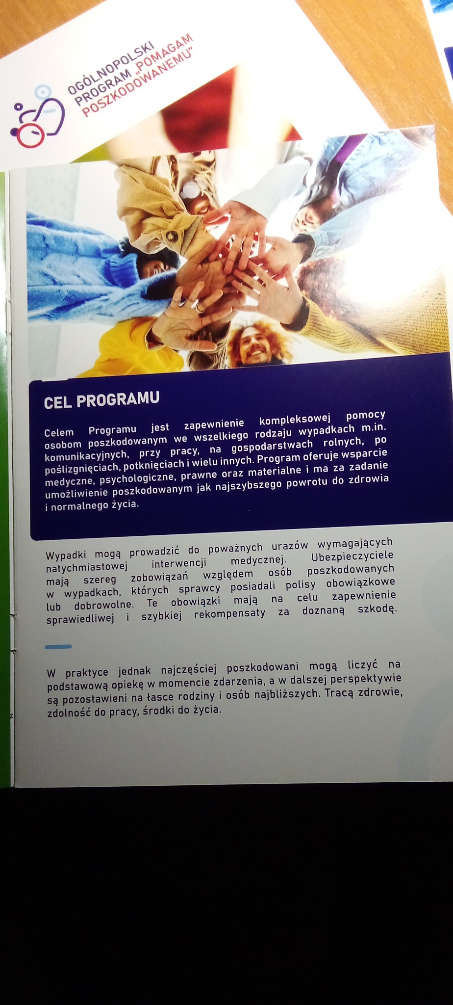 Ulotka informacyjna programu 'Pomagam Poszkodowanemu' z ilustracją dłoni złączonych w geście wsparcia i tekstem o celach programu.