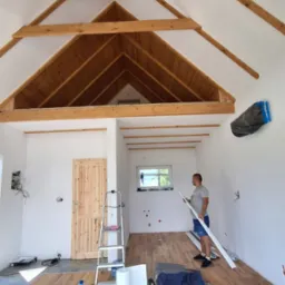 Wnętrze domu w trakcie wykańczania: drewniana konstrukcja dachu, białe ściany, podłoga imitująca drewno, mężczyzna trzymający białą listwę przypodłogową, widoczne narzędzia i materiały budowlane.