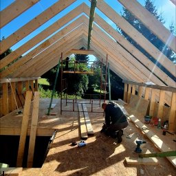 PAWBUD Paweł Będkowski - Konstrukcja drewniana dachu w trakcie budowy domu szkieletowego, widok z wnętrza. Pracownik w trakcie pracy, narzędzia budowlane w pobliżu. Słoneczny dzień.