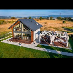 PAWBUD Paweł Będkowski - Nowoczesny dom szkieletowy z dużymi przeszkleniami i pergolą na tarasie, otoczony zielonym trawnikiem i polami. Widok z lotu ptaka, słoneczny dzień.