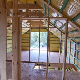 PAWBUD Paweł Będkowski - Wnętrze domu szkieletowego w trakcie budowy: drewniana konstrukcja ścian i dachu, żółta folia izolacyjna, widok na zewnątrz przez otwór okienny. Podłoga z płyt OSB.