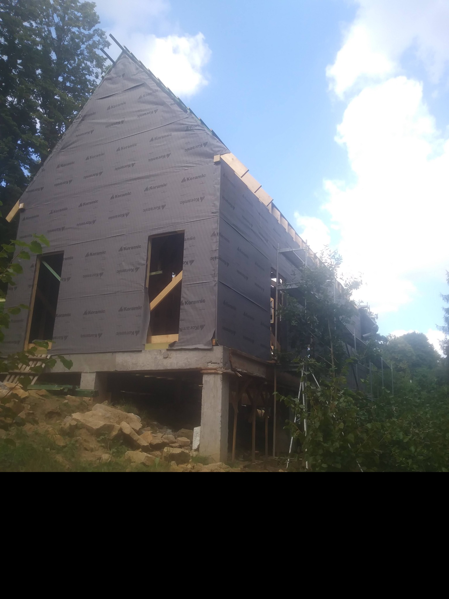 Surowy stan domu szkieletowego w lesistej okolicy, pokrytego membraną ochronną marki Koramic, z widocznymi drewnianymi elementami konstrukcyjnymi i fragmentem rusztowania.