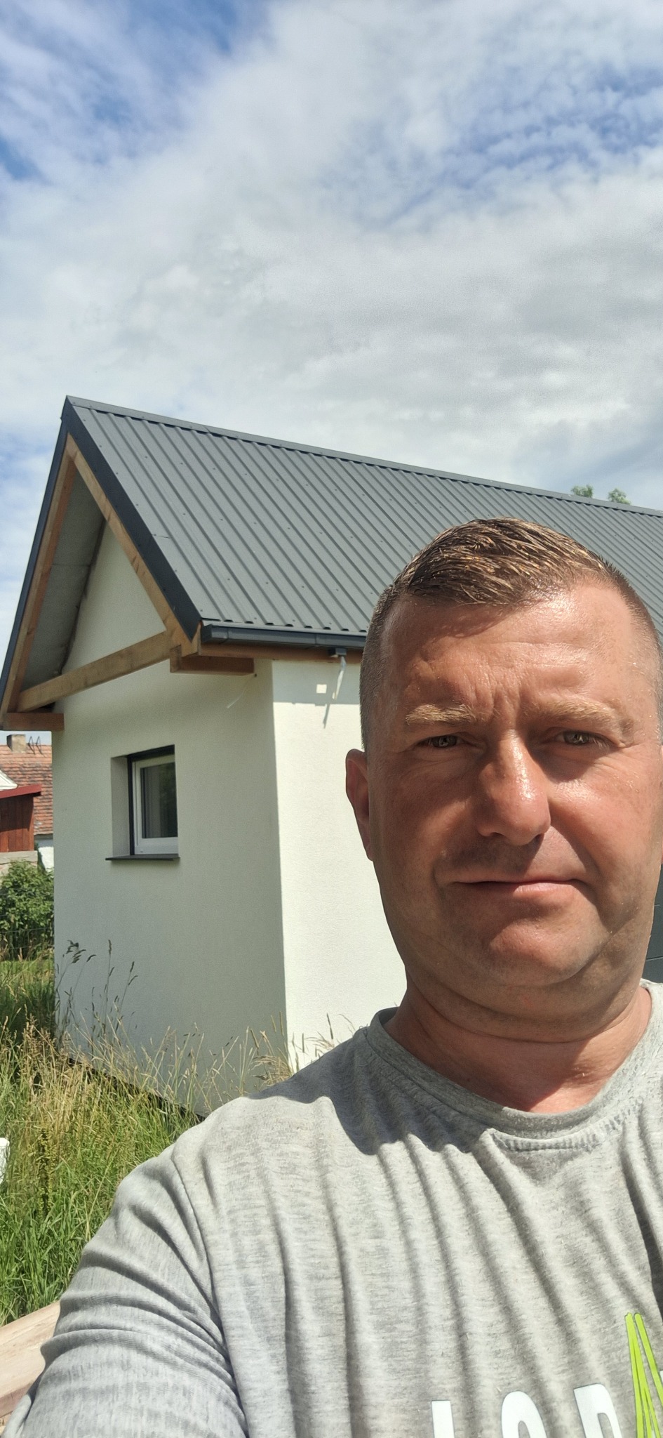 Mężczyzna robiący selfie na tle nowo wybudowanego, otynkowanego domu z szarą blachodachówką i drewnianą konstrukcją dachu widoczną spod okapu.