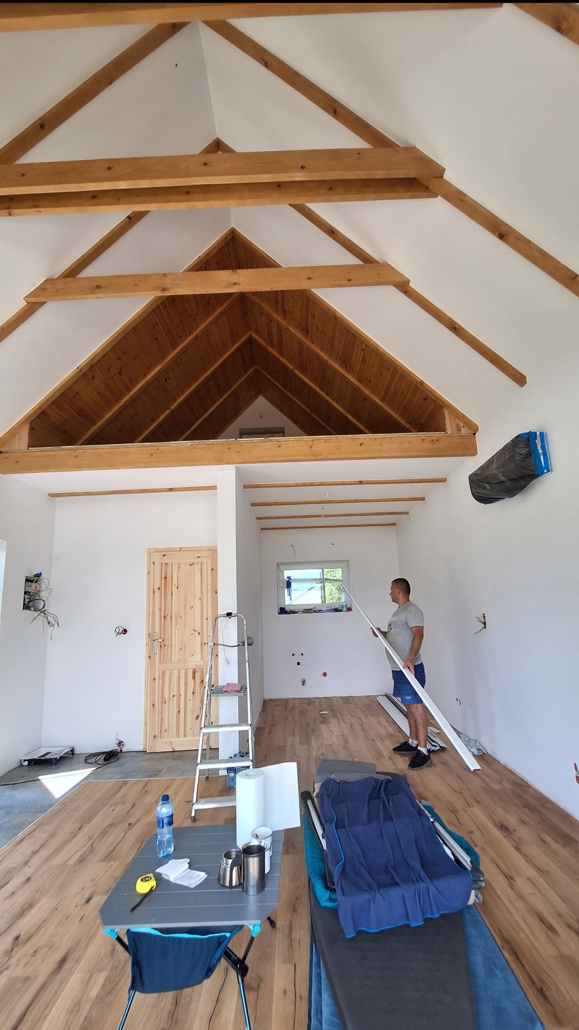 Wnętrze domu w trakcie wykańczania: drewniana konstrukcja dachu, białe ściany, podłoga imitująca drewno, mężczyzna trzymający białą listwę przypodłogową, widoczne narzędzia i materiały budowlane.
