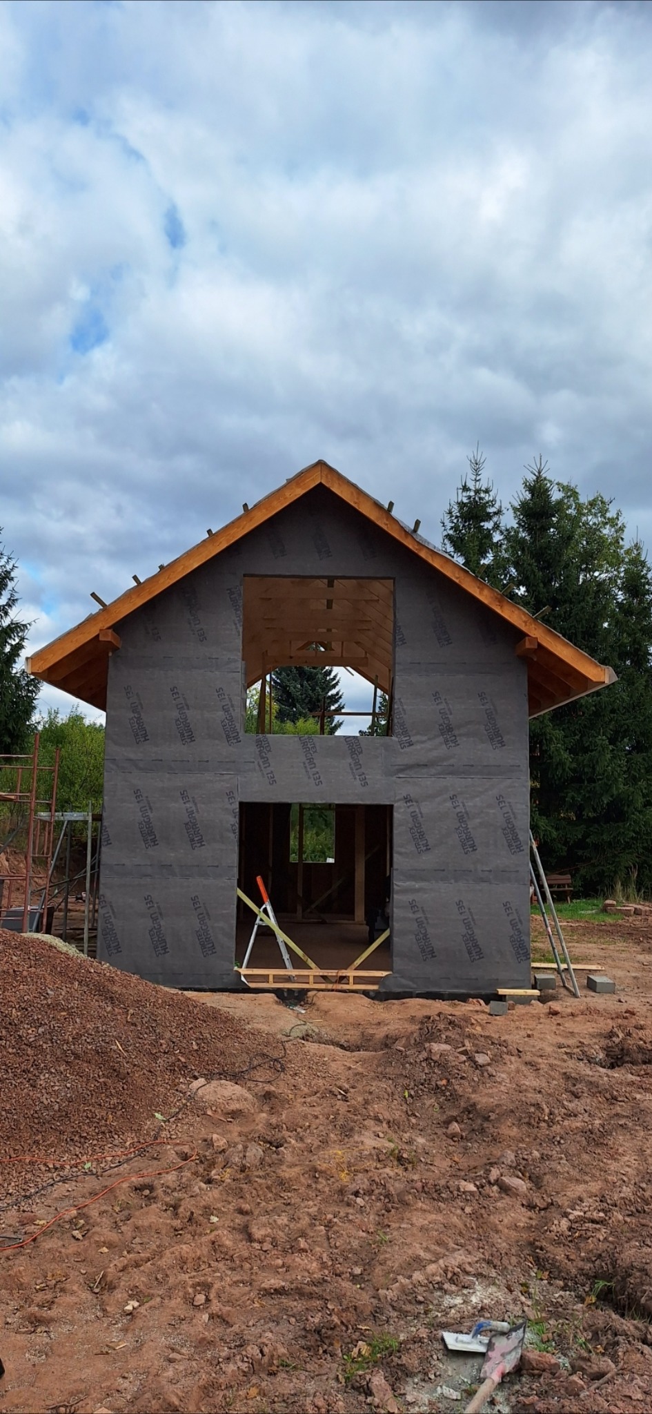 Surowy stan domu szkieletowego z membraną ochronną na ścianach, widoczne drewniane elementy konstrukcyjne i narzędzia budowlane. W tle drzewa i pochmurne niebo.