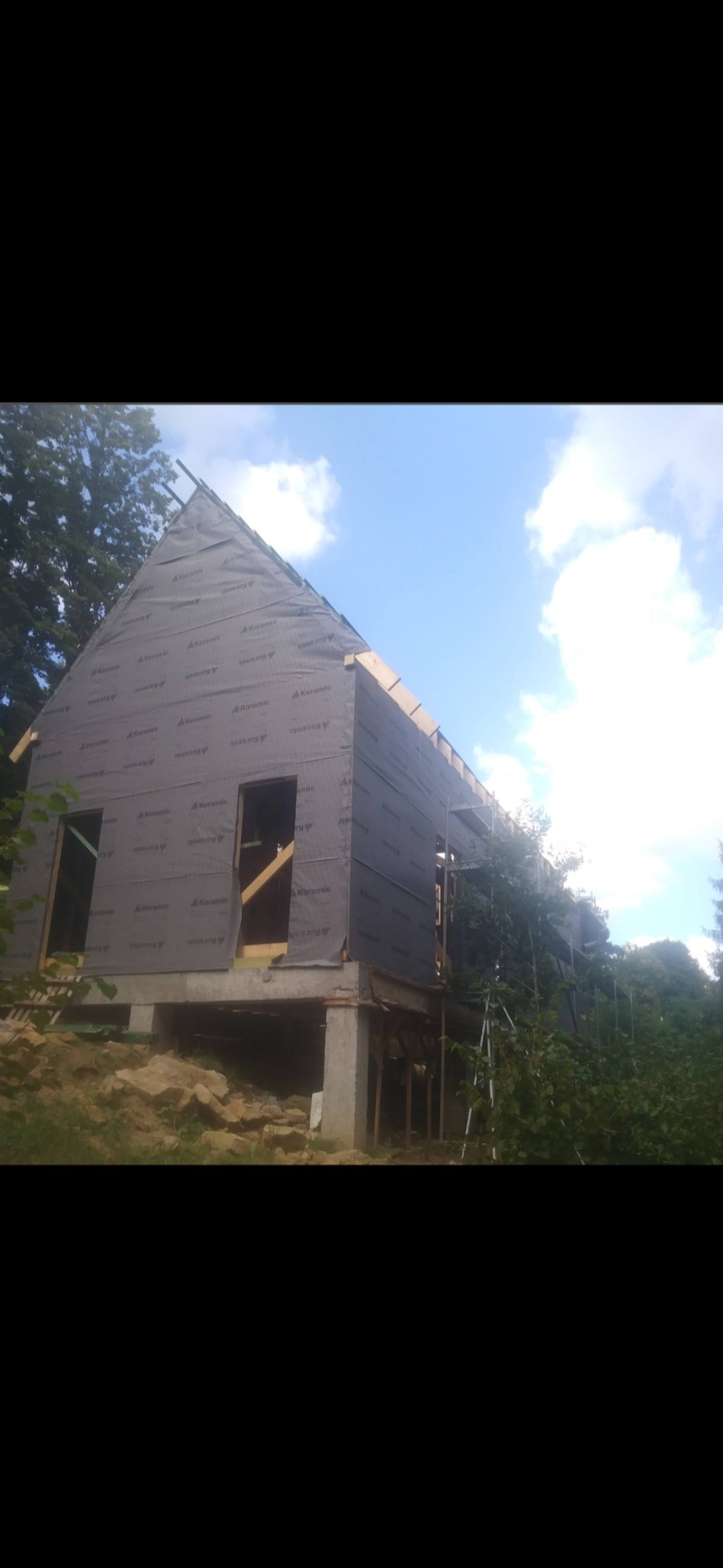 Budowa domu szkieletowego w lesie: widok na konstrukcję z membraną, okna i drzwi w trakcie montażu, betonowe fundamenty. Prace budowlane w toku, drzewa w tle.