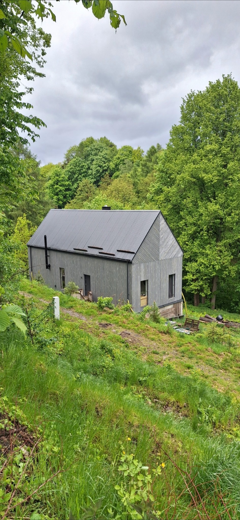 Nowoczesny dom szkieletowy z ciemną blachą na dachu, otoczony zielenią w Kłodzku. Widok z dołu, podkreślający naturalne otoczenie i minimalistyczny design.