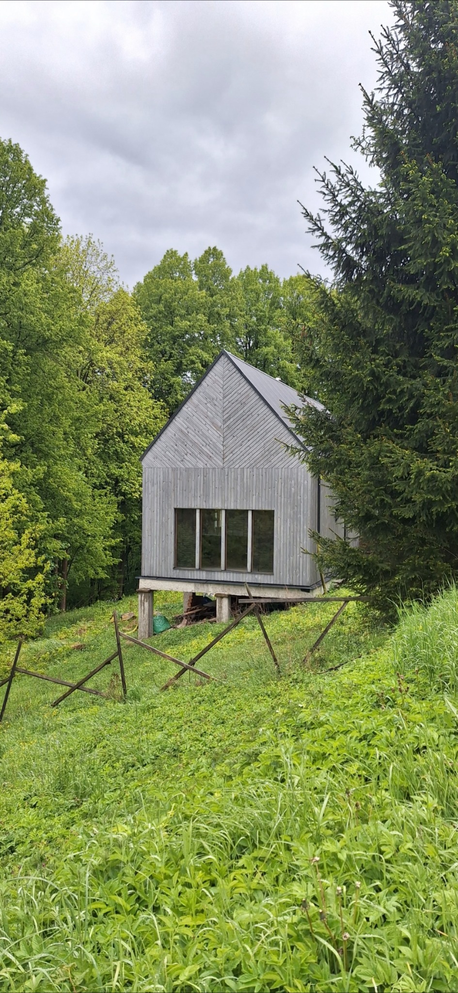 Nowoczesny dom szkieletowy z szarego drewna, wkomponowany w zielony krajobraz. Widoczne trzy okna i drewniane wykończenie elewacji. Dom na wzniesieniu.