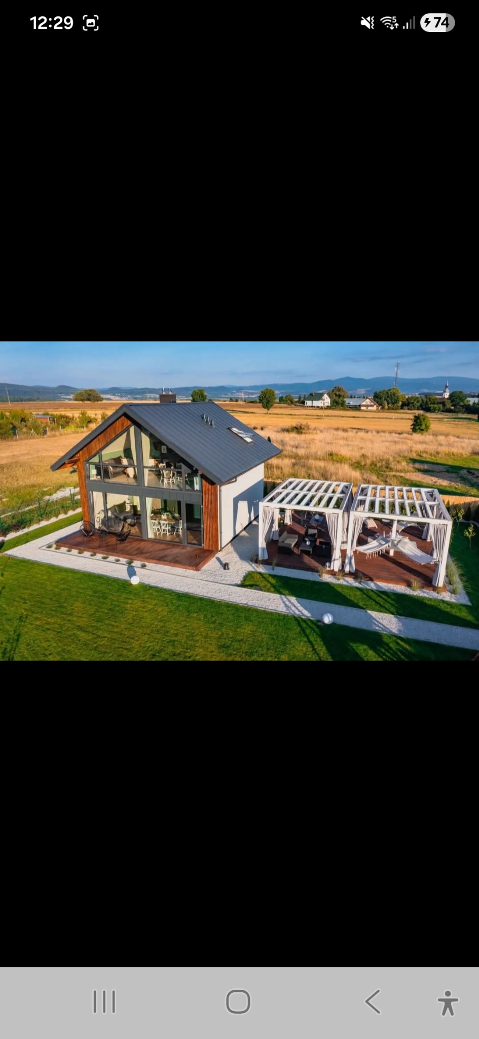 Nowoczesny dom szkieletowy z dużymi przeszkleniami i pergolą na tarasie, otoczony zielonym trawnikiem i polami. Widok z lotu ptaka, słoneczny dzień.