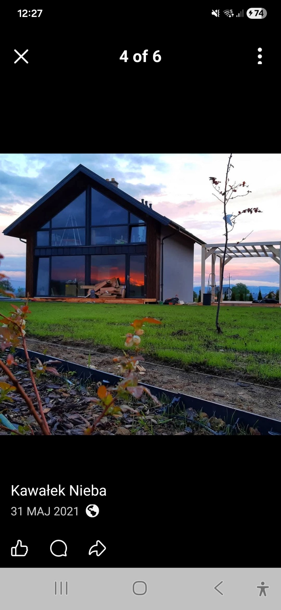 Nowoczesny dom szkieletowy z dużymi oknami, odbijającymi zachód słońca, otoczony zielonym trawnikiem i młodymi roślinami. Widoczna drewniana pergola.