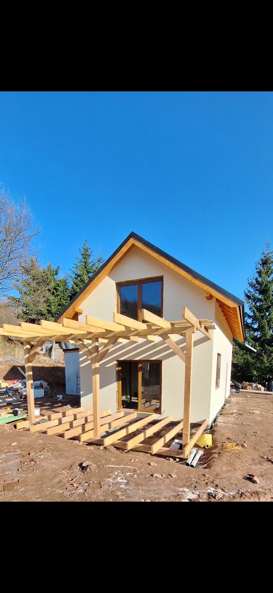 Niedokończony dom szkieletowy z drewnianą konstrukcją tarasu na ziemnym podłożu, widok z zewnątrz w słoneczny dzień. Widoczne okna i drzwi balkonowe.