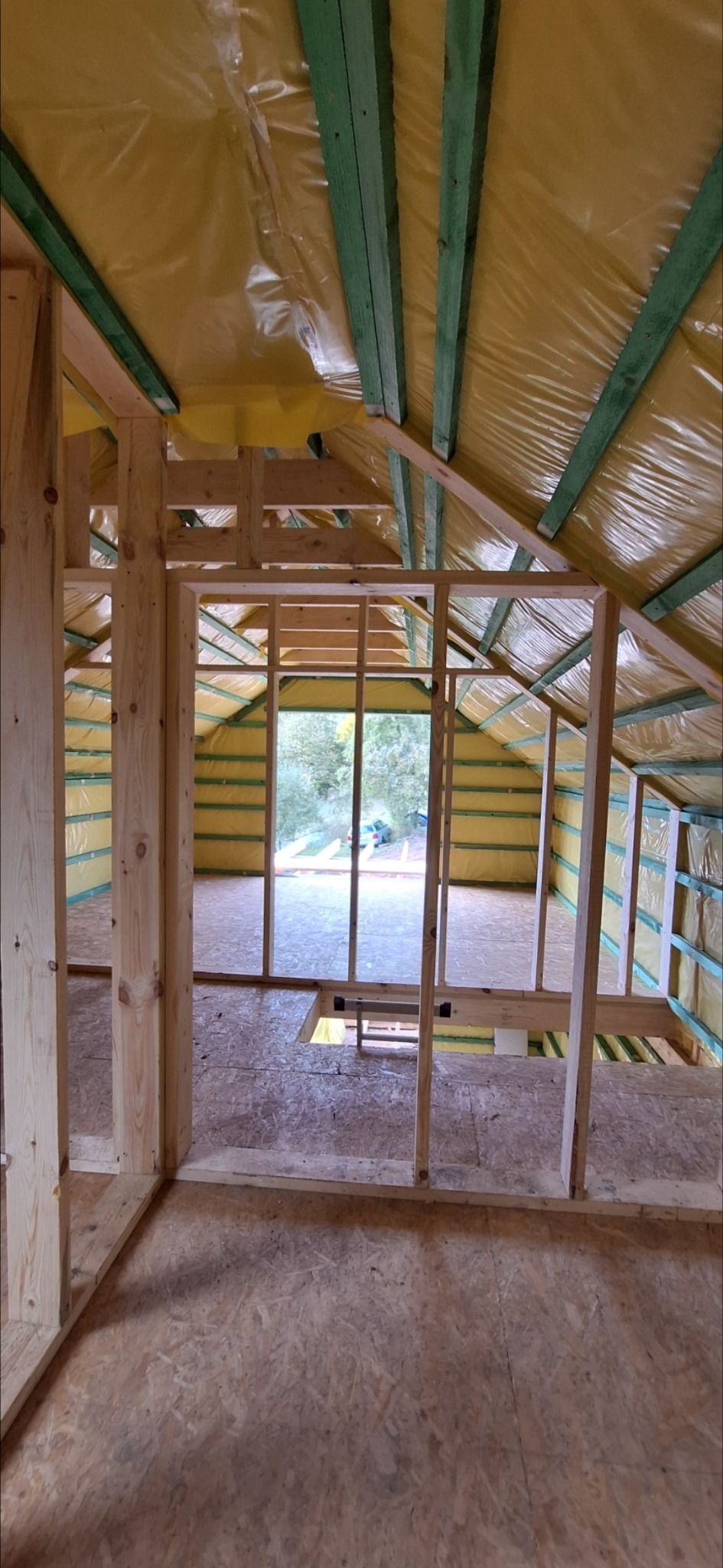 Wnętrze domu szkieletowego w trakcie budowy: drewniana konstrukcja ścian i dachu, żółta folia izolacyjna, widok na zewnątrz przez otwór okienny. Podłoga z płyt OSB.