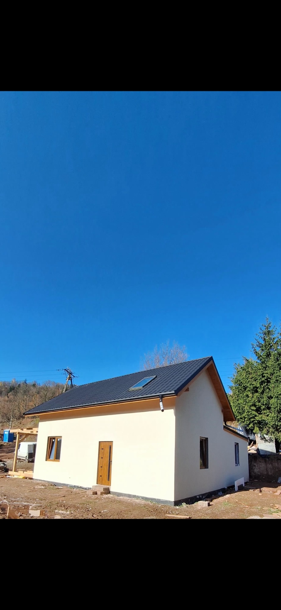 Nowo wybudowany, parterowy dom z jasną elewacją, ciemnym dachem i drewnianymi oknami. Widoczne elementy konstrukcyjne i otoczenie w trakcie zagospodarowania.