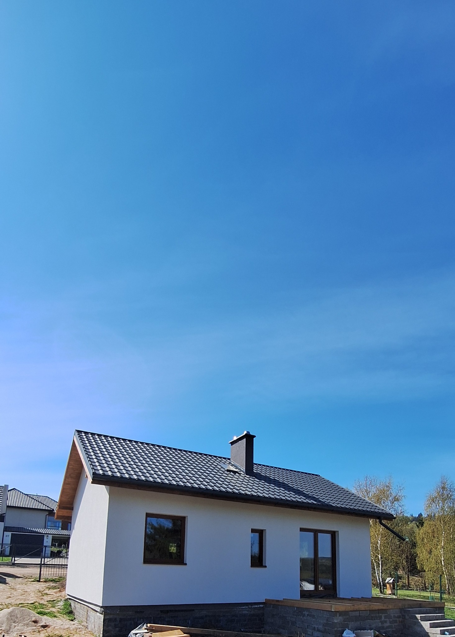 Biały dom z ciemną dachówką i kominem na tle błękitnego nieba. Widoczne okna z brązowymi ramami i częściowo zadaszony taras. W tle drzewa i inne domy.
