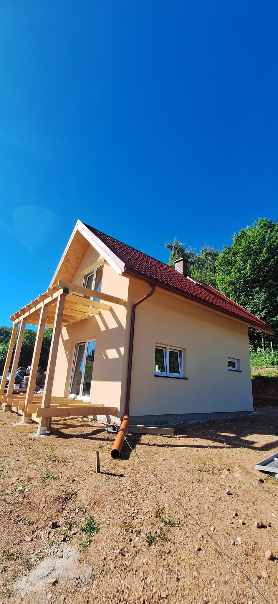 Nowo wybudowany dom jednorodzinny z jasnobeżową elewacją, czerwoną dachówką i częściowo ukończoną drewnianą pergolą tarasową, otoczony niezagospodarowanym terenem.