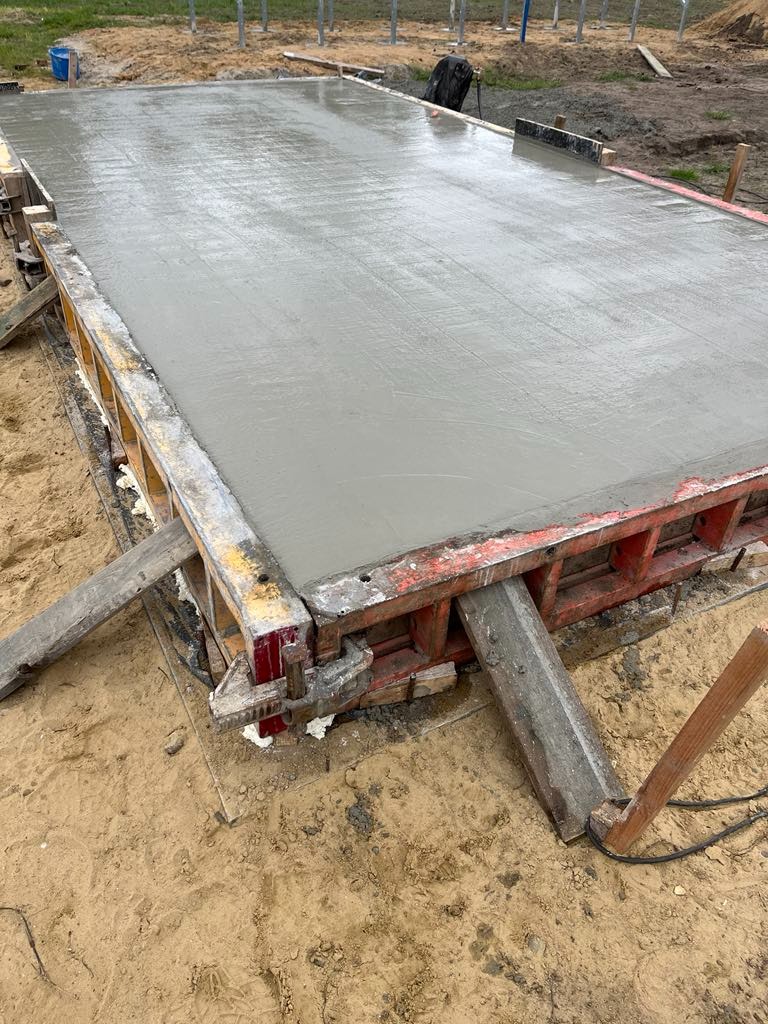 Świeżo wylana, gładka płyta betonowa w szalunku z czerwonymi elementami, podparta drewnianymi stemplami, na piaszczystym podłożu.