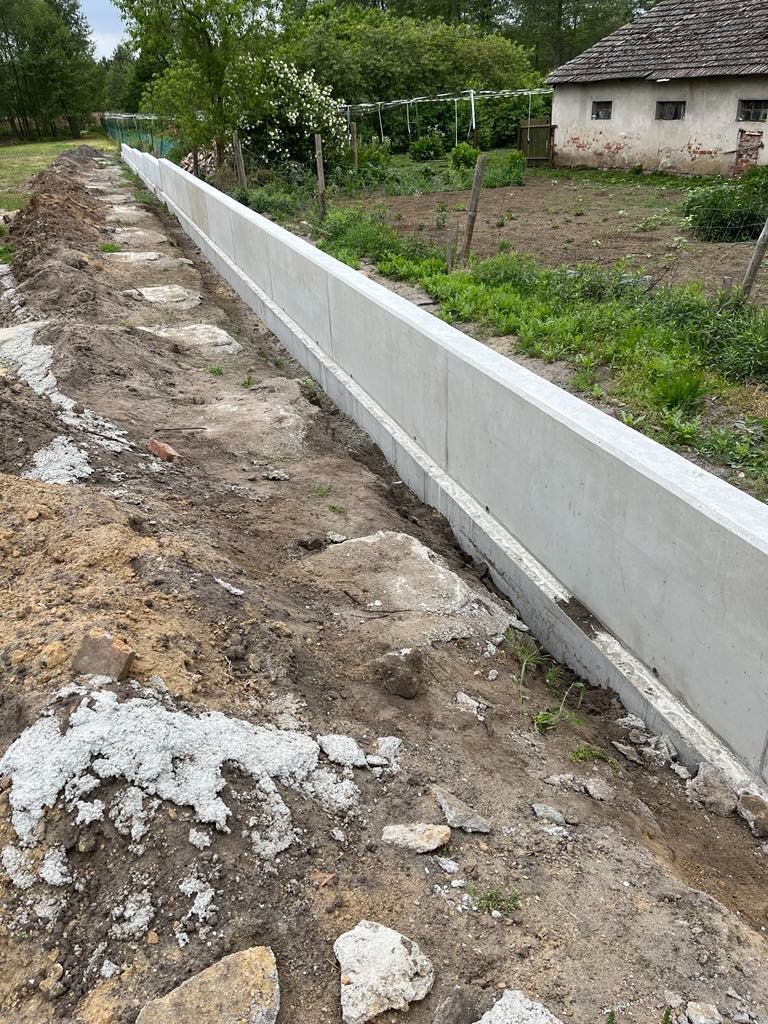 Długi, szary, betonowy mur oporowy w trakcie budowy, widoczny w krajobrazie z ziemią i zielenią, z domem w tle.