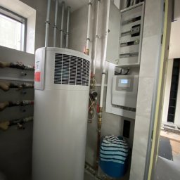 MOMA MONIKA TOKARSKA-PASTERNAK - Biała pompa ciepła z widocznymi rurami i instalacją elektryczną w jasnym pomieszczeniu podczas montażu.