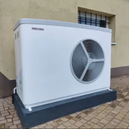 Biała pompa ciepła Stiebel Eltron zamontowana na betonowym cokole przed budynkiem z oknem zabezpieczonym kratą.