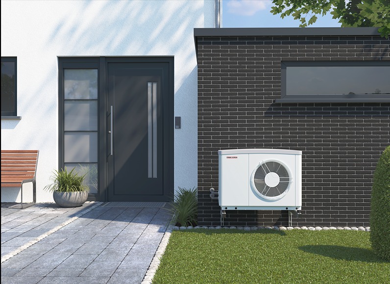 Biała pompa ciepła zamontowana na zewnątrz budynku z ciemnej cegły, obok wejścia z szarymi drzwiami i chodnikiem z kostki.
