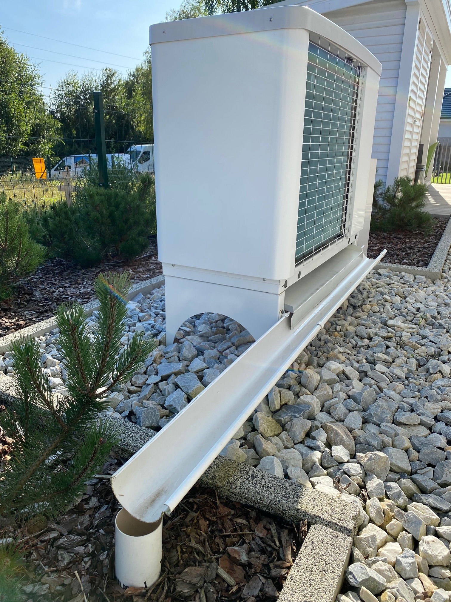 Biała pompa ciepła typu monoblok z odprowadzeniem kondensatu, zainstalowana na posesji z ozdobnymi kamieniami i zielenią.