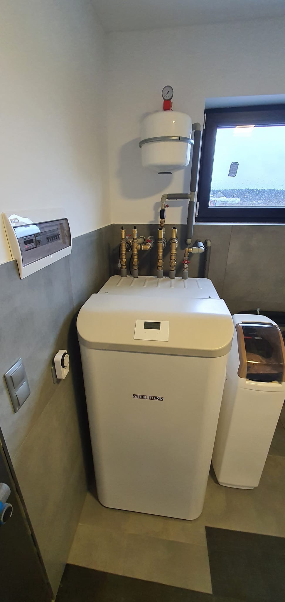 Biała pompa ciepła Stiebel Eltron z widocznymi przyłączami hydraulicznymi, zbiornikiem wyrównawczym i panelem elektrycznym w szarej kotłowni.