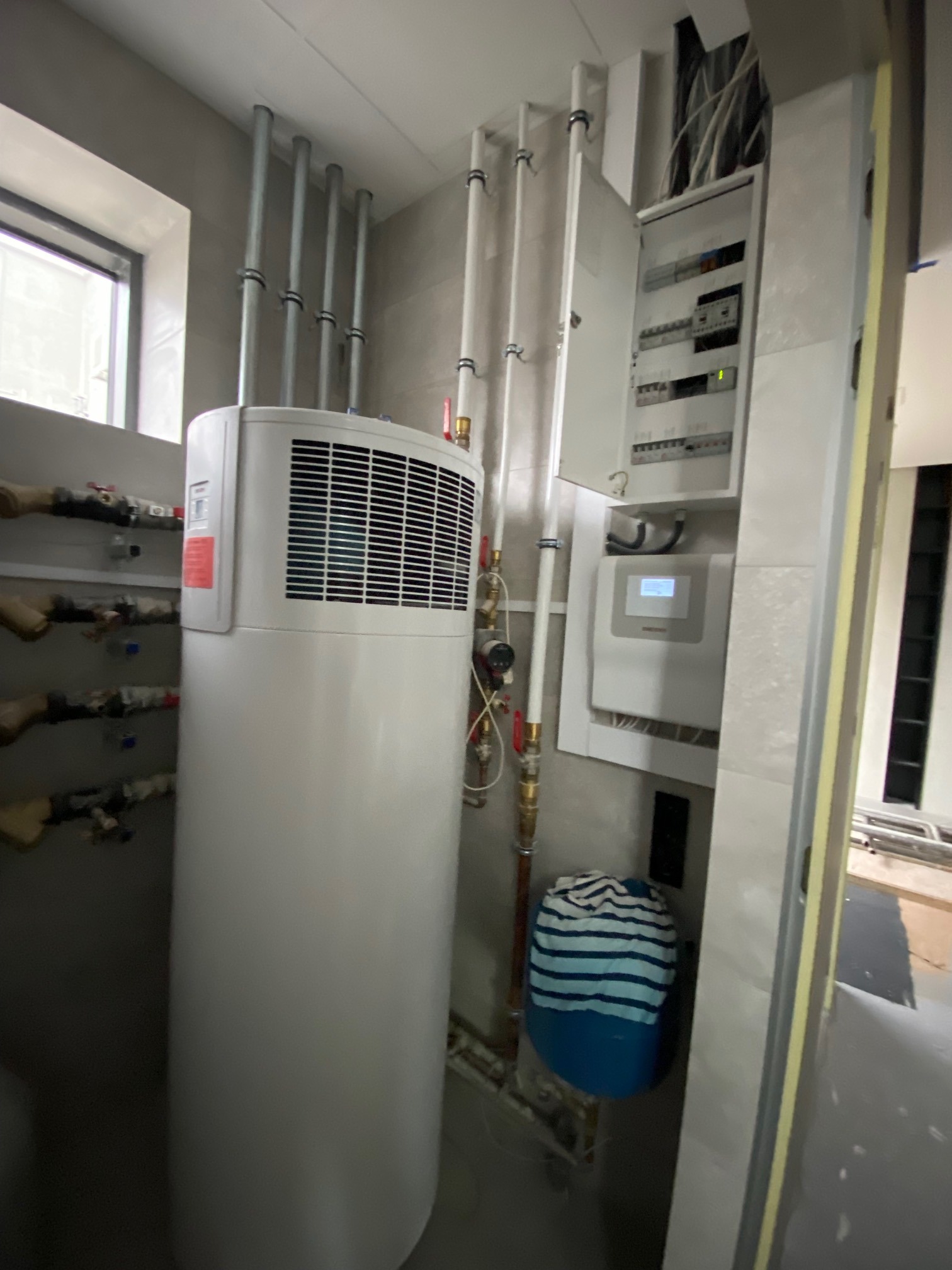 Biała pompa ciepła z widocznymi rurami i instalacją elektryczną w jasnym pomieszczeniu podczas montażu.