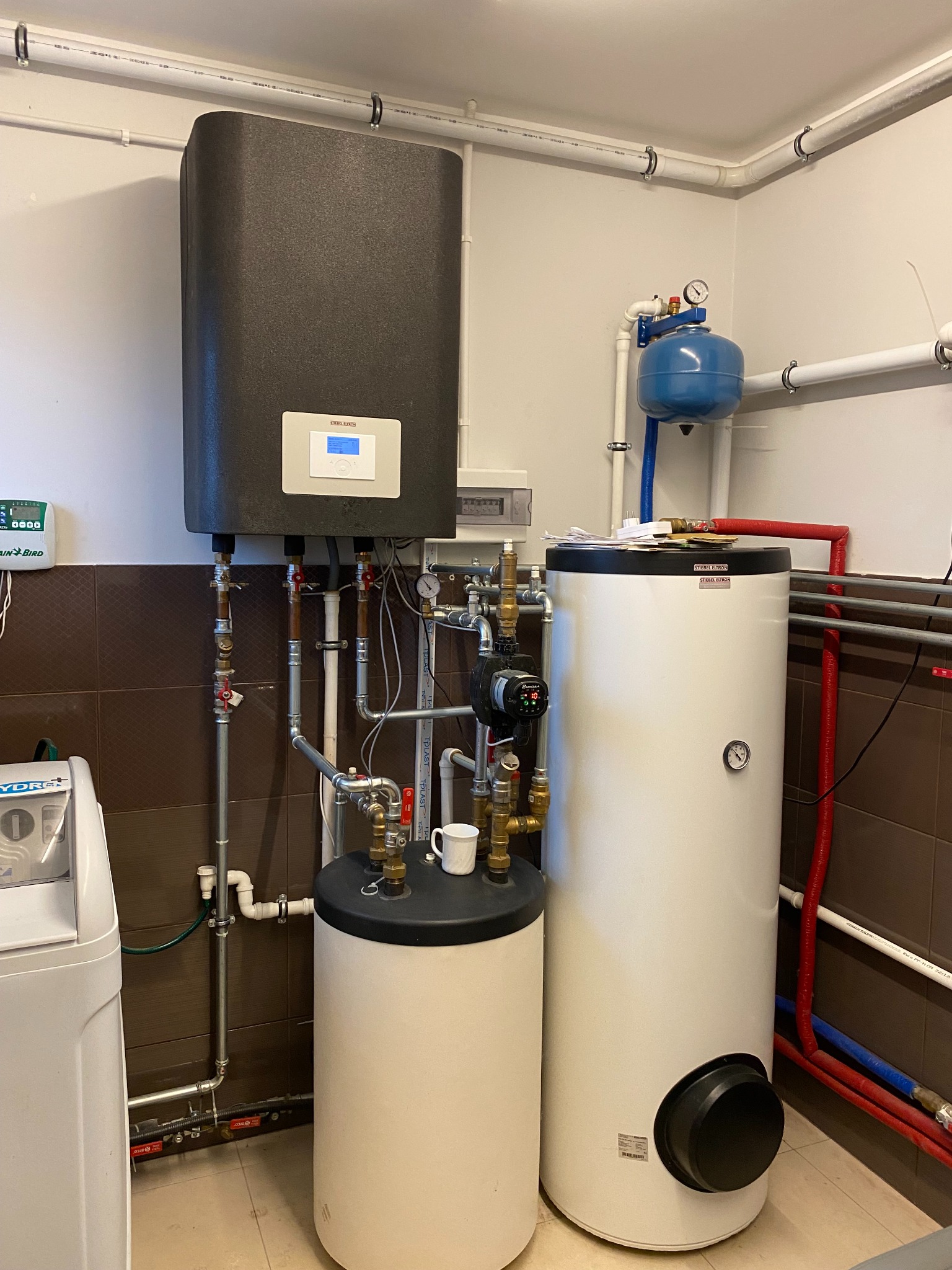 Kompletna instalacja grzewcza w kotłowni: pompa ciepła z zasobnikiem, zbiornik wyrównawczy i instalacja wodna. Na zasobniku stoi biały kubek.