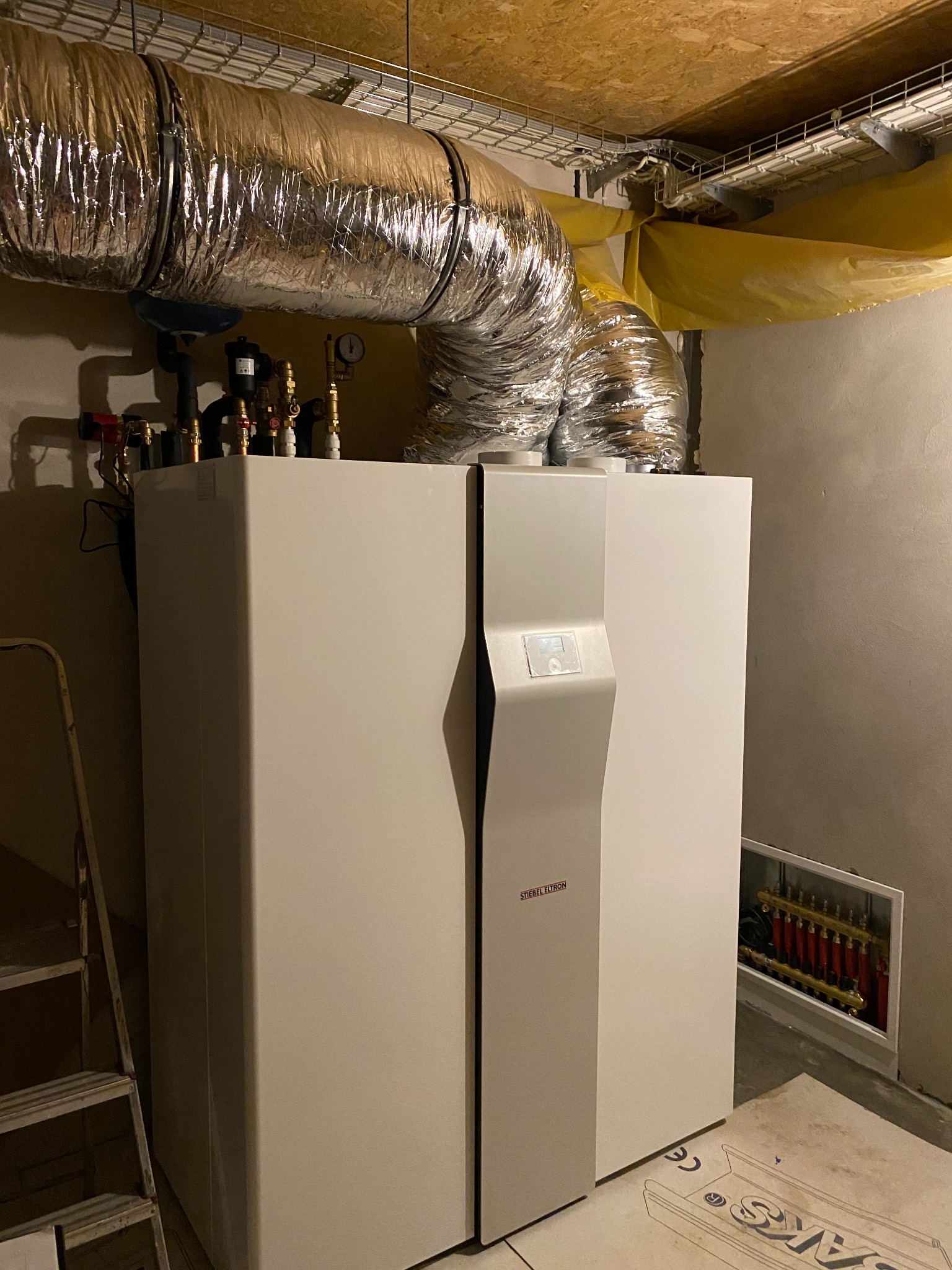 Biała pompa ciepła Stiebel Eltron z podłączonymi rurami i przewodami wentylacyjnymi w pomieszczeniu technicznym, widoczna drabina i rozdzielacz ogrzewania podłogowego.