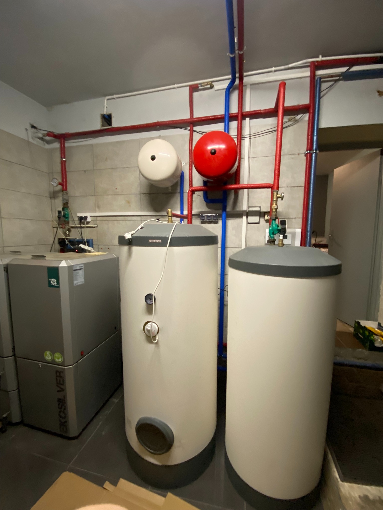 Kotłownia z dwoma zasobnikami ciepłej wody użytkowej marki Stiebel Eltron, kotłem EkoSilver, oraz instalacją hydrauliczną z czerwonymi i niebieskimi rurami.