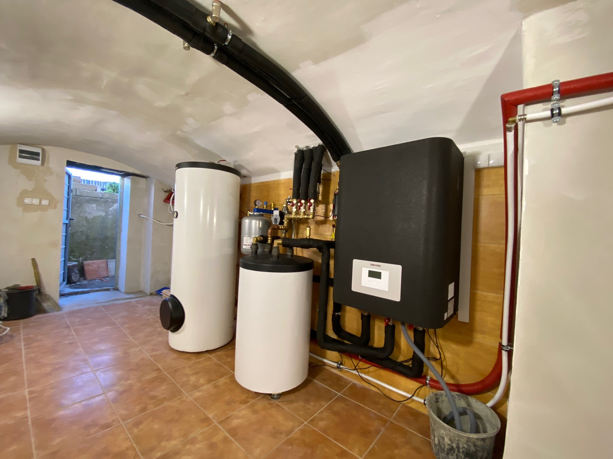 Instalacja grzewcza w piwnicy z cylindrycznymi zbiornikami i czarnym panelem sterującym, widoczne rury i armatura, surowe wykończenie wnętrza.
