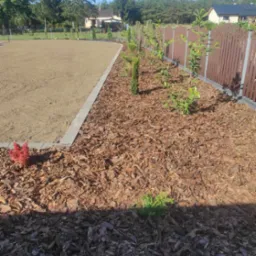 Przygotowany teren pod trawnik z obrzeżem i świeżo posadzone drzewka wzdłuż ogrodzenia, obsypane korą.