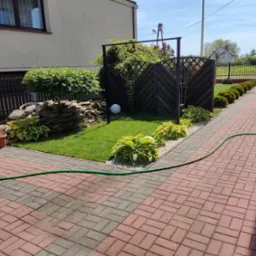 Uporządkowany ogród z trawnikiem, rabatami host, kamienną kaskadą, drewnianą pergolą i zielonym wężem ogrodowym na kostce brukowej.