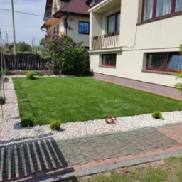 Widok na równo przystrzyżony trawnik, obrzeża z białych kamieni, ozdobne krzewy i drzewka w ogrodzie przed domem z jasną elewacją.