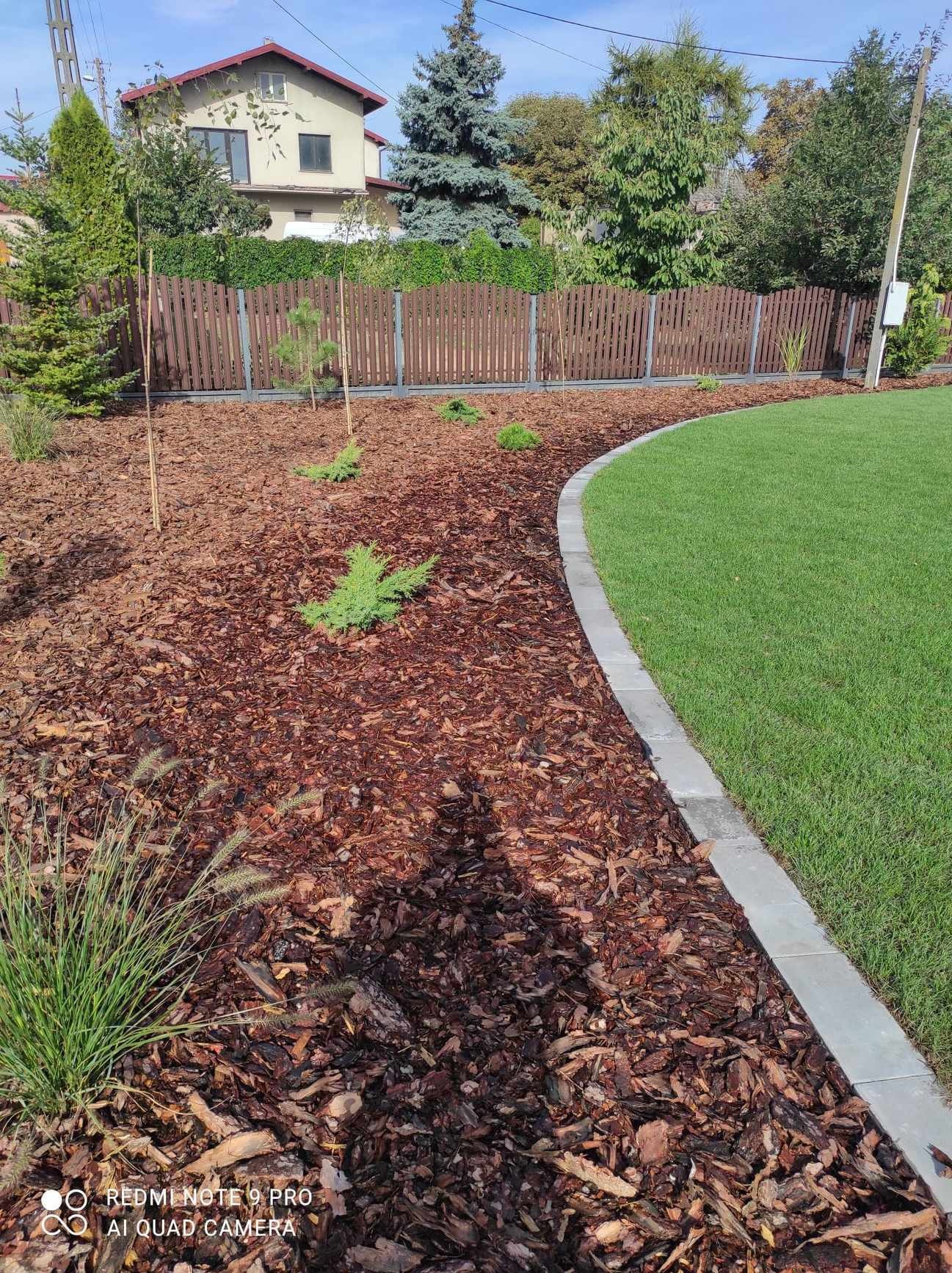 Świeżo założony ogród z korą, młodymi drzewkami i równo przystrzyżonym trawnikiem oddzielonym betonowym obrzeżem, widok na brązowe ogrodzenie i dom w tle.