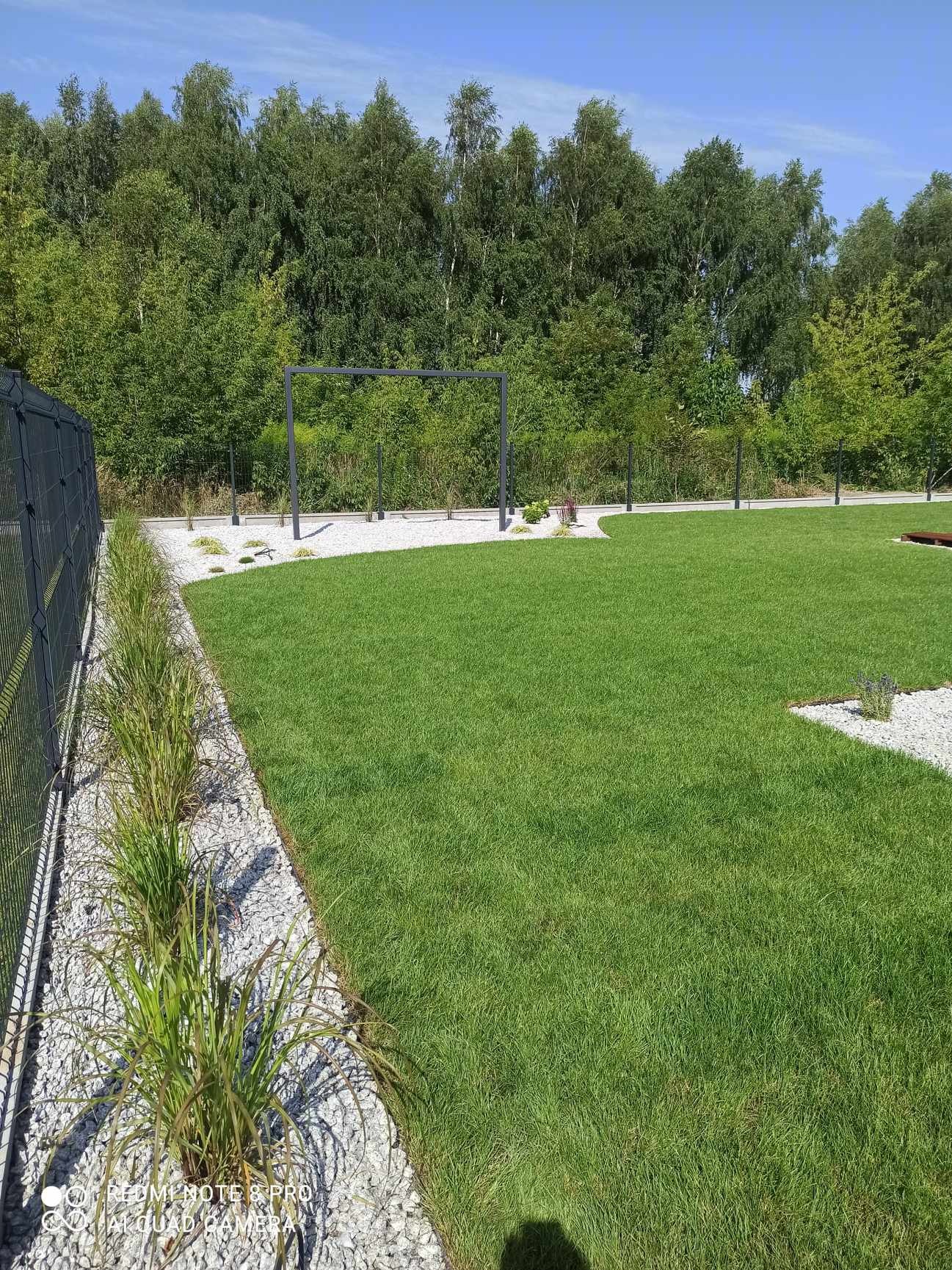 Starannie utrzymany trawnik z równo przystrzyżoną trawą, obrzeżami z białego kamienia, dekoracyjnymi trawami wzdłuż ogrodzenia i minimalistyczną pergolą w tle.