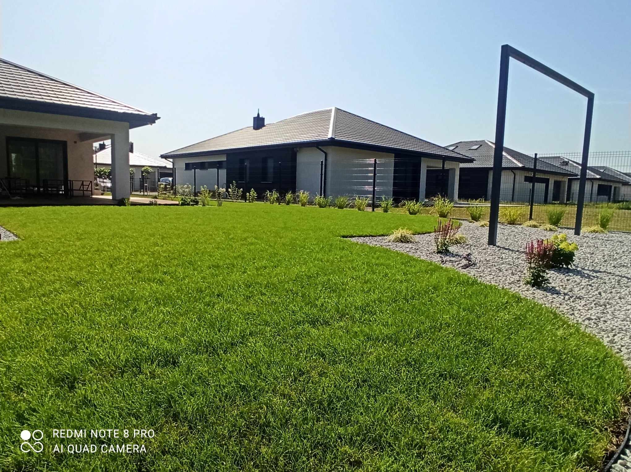 Świeżo założony trawnik z rolki przed nowoczesnym domem jednorodzinnym, widoczne obrzeża trawnika i rabaty wysypane drobnym żwirem, w tle kolejne domy.