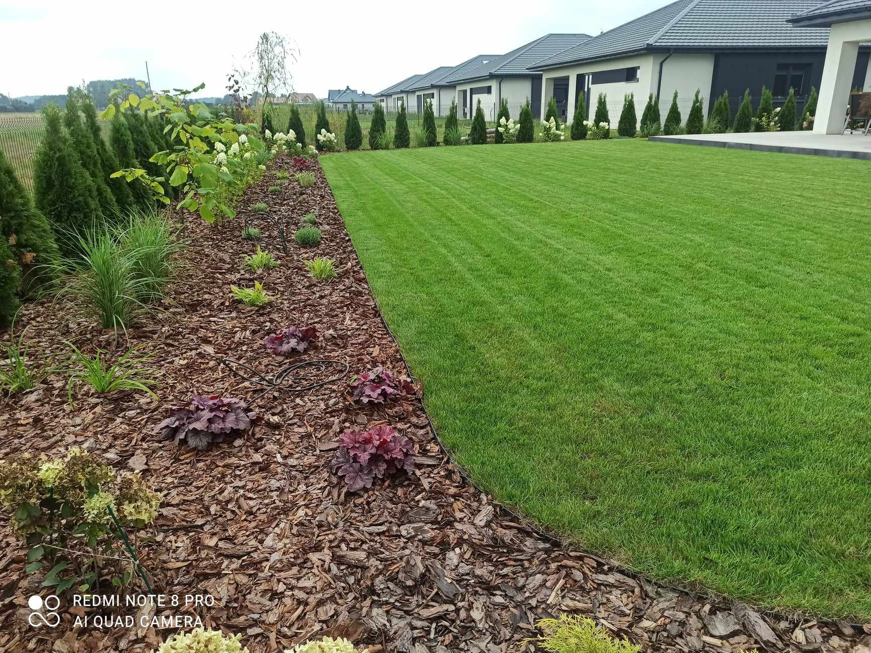 Perfekcyjnie utrzymany trawnik z równo przystrzyżoną trawą, obrzeże z kory i nasadzeniami ozdobnymi, w tle nowoczesne domy jednorodzinne.